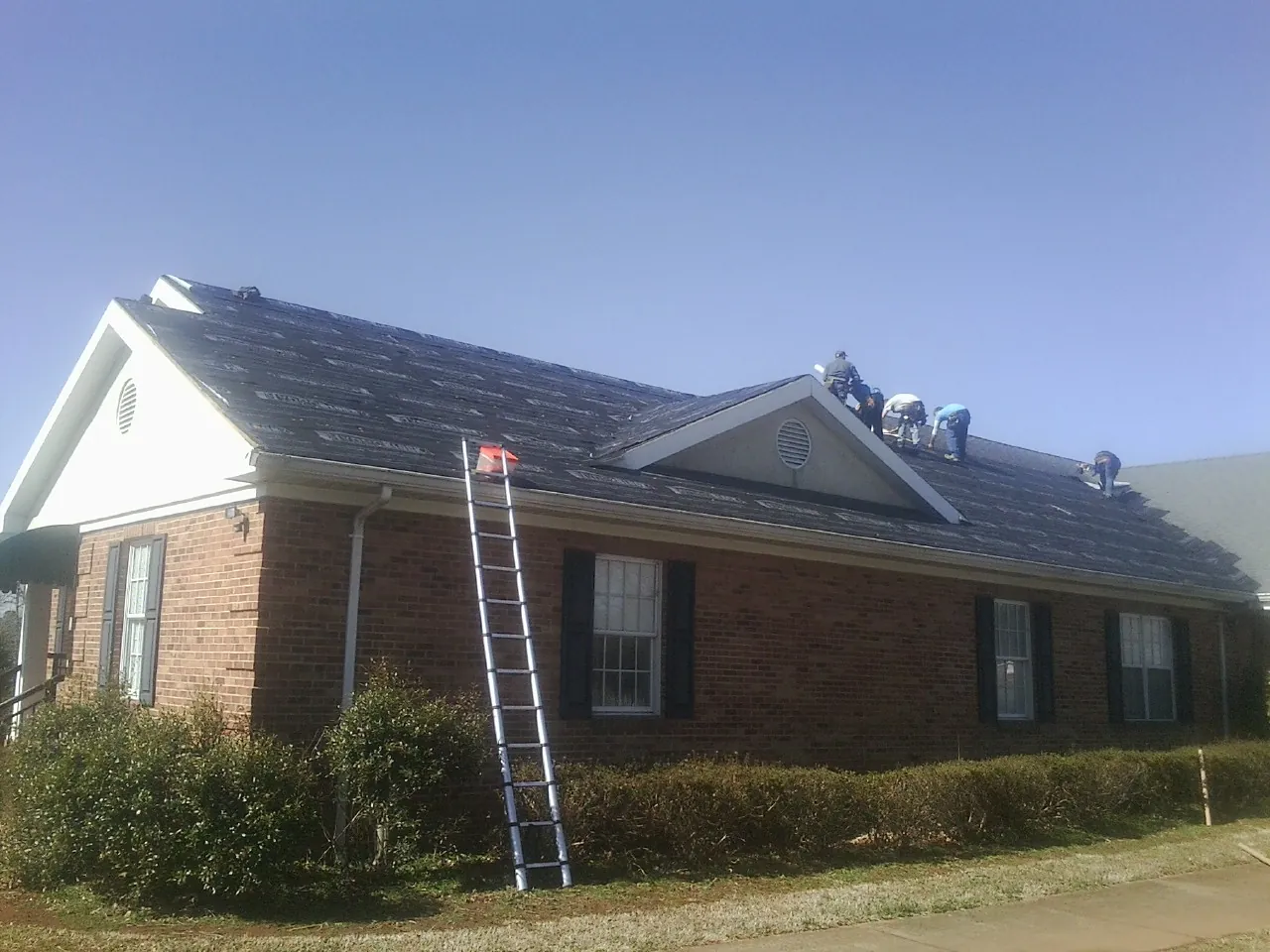 Crew tearing shingles off a brick ranch roof with a ladder set — Metal Roof Repair work by our crew in Steamboat Rock