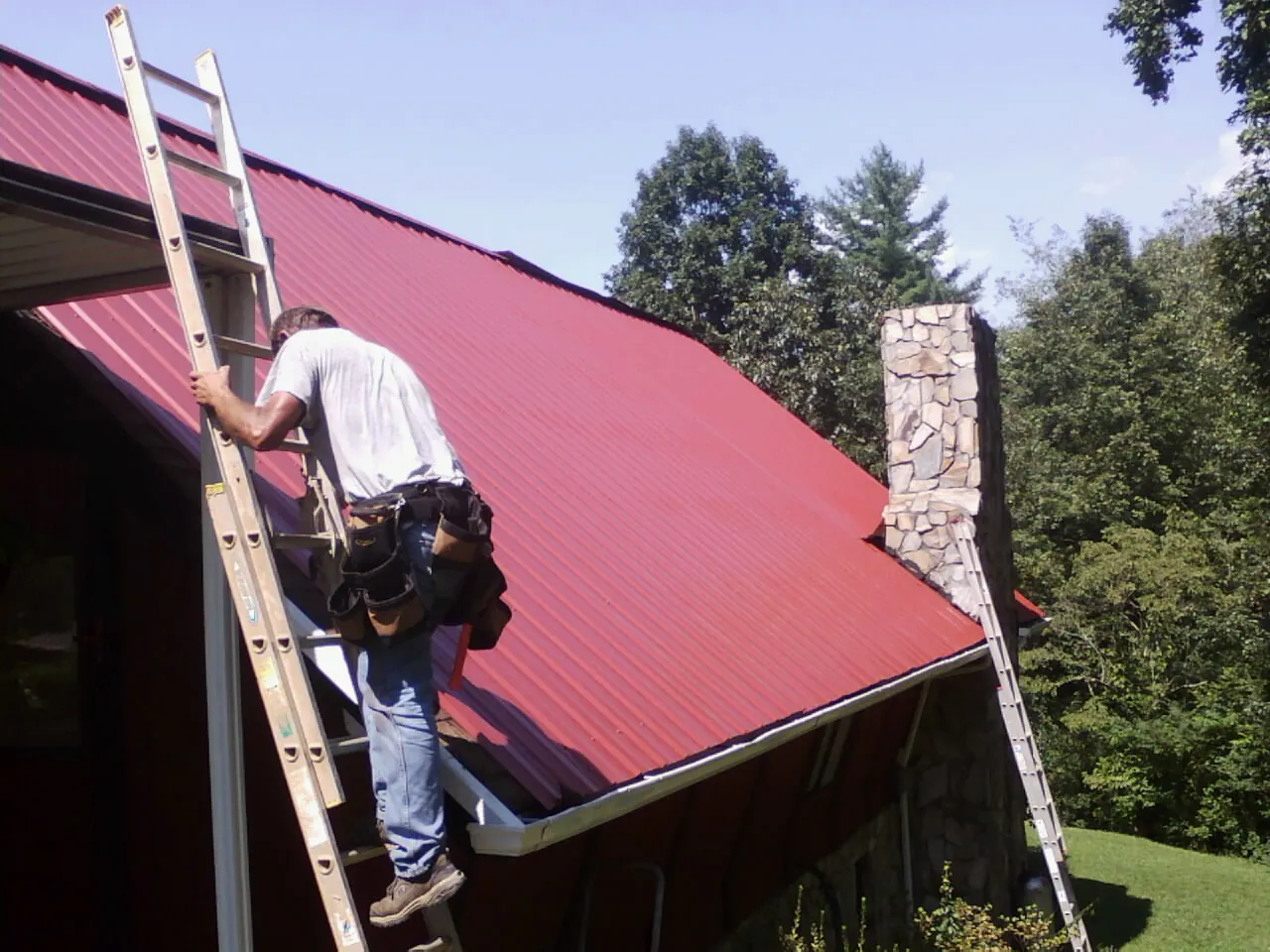 Roofer on a ladder beside a red metal roof and stone chimney — Soffit & Fascia Repair by Roofing Brothers in Steamboat Rock
